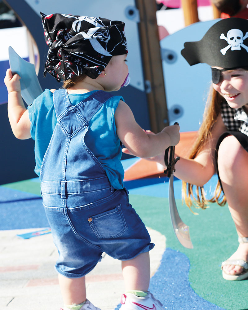 A young toddler walks towards an older child, they are both dressed up pirates at a pirate themed playground.